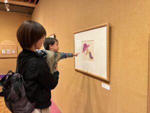 ファーストミュージアムデー　子どもを抱えた大人が作品の前に立っている。子どもは作品に手を伸ばして指をさしている。